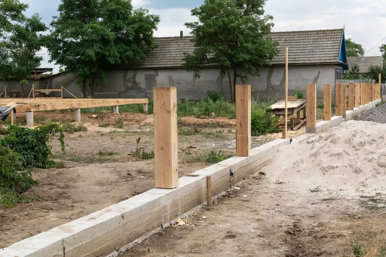Budowa ogrodzenia: betonowy fundament i drewniane słupki w trakcie prac. W tle widoczna konstrukcja podłogi na palach.