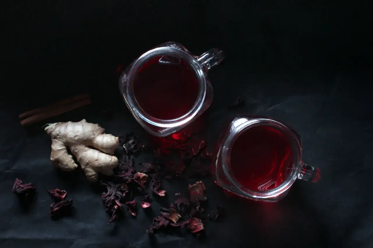 Deux verres de bissap chaud, accompagn&eacute;s de gingembre et de b&acirc;tons de cannelle, sur fond sombre.