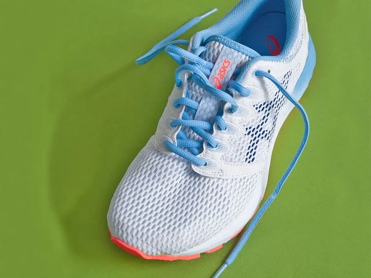 Ein weißer Laufschuh mit hellblauen Schnürsenkeln, bereit zum Schnüren, liegt auf einem leuchtend grünen Hintergrund.