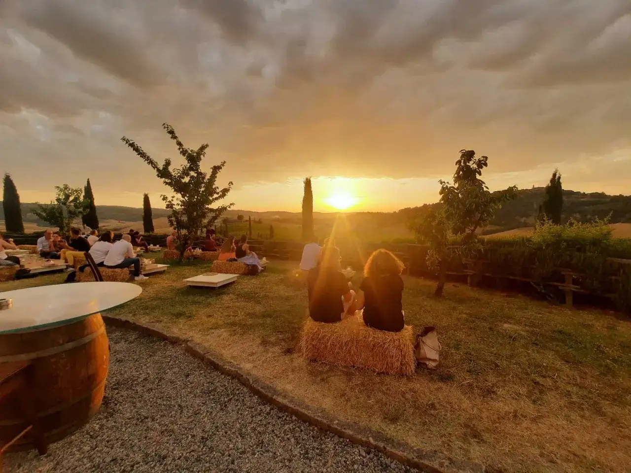 Tramonto in Toscana con degustazione vini. Persone sedute su balle di fieno godono del paesaggio collinare.