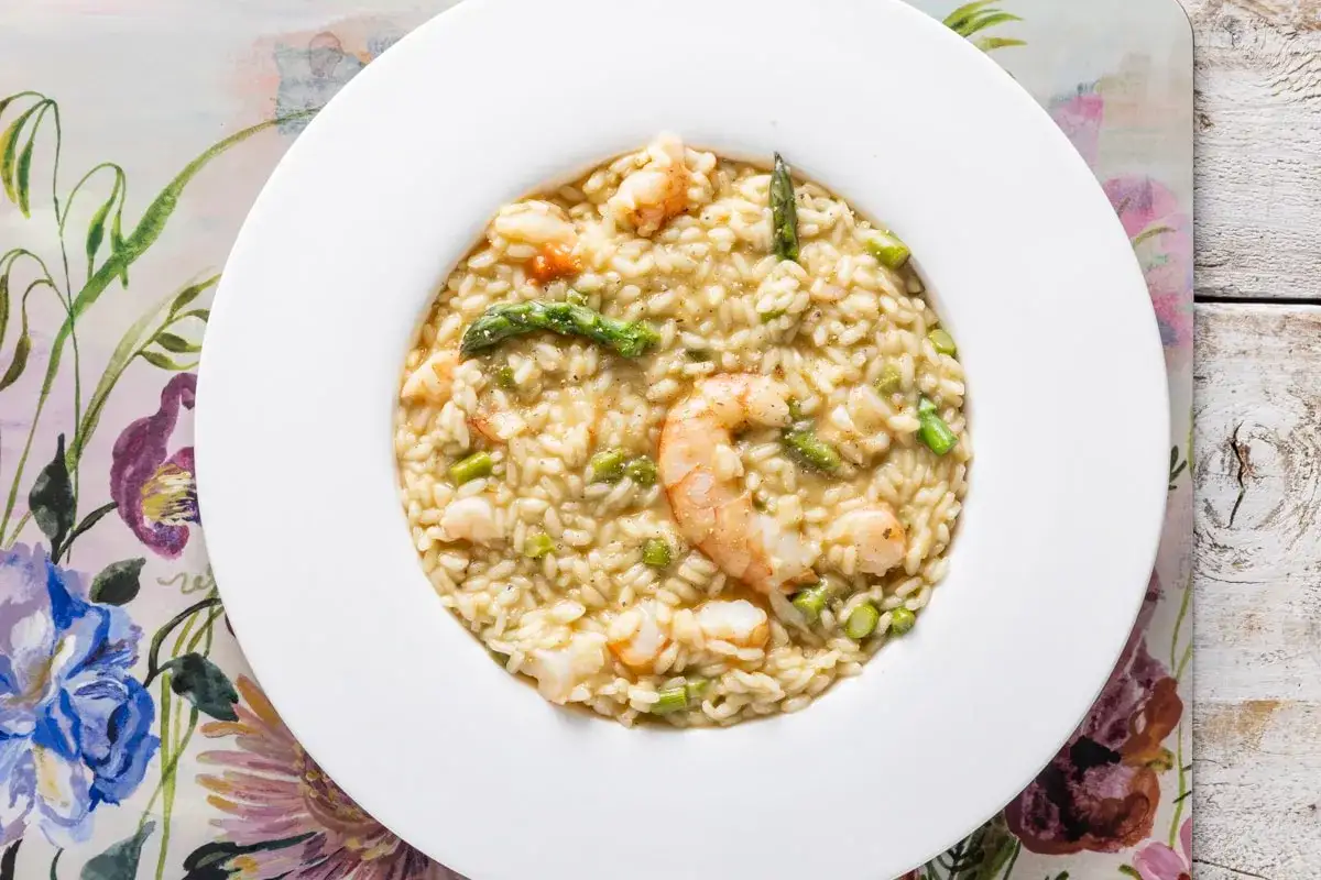 Un cremoso risotto asparagi e gamberetti, con gamberi succulenti e punte di asparagi verdi, servito in un piatto bianco su un tovagliolo fiorito.