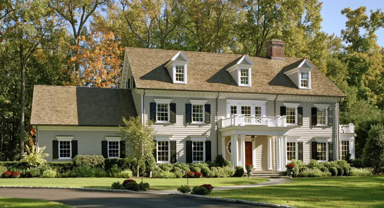 A beautiful home showcasing classic colonial architecture, with symmetrical windows, a prominent portico, and dormers, nestled amongst vibrant autumn trees.