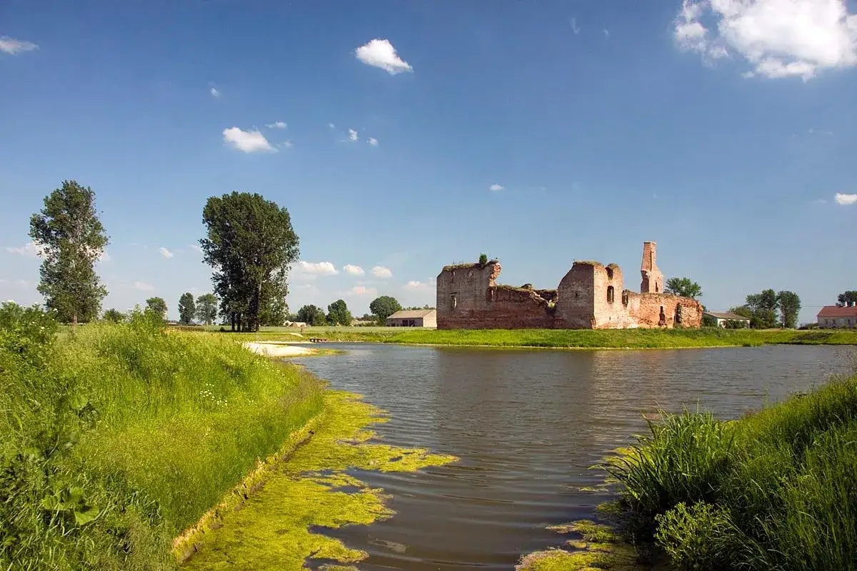 Ruiny zamku w Besiekierach nad spokojną wodą, otoczone zielenią i błękitnym niebem z chmurami.