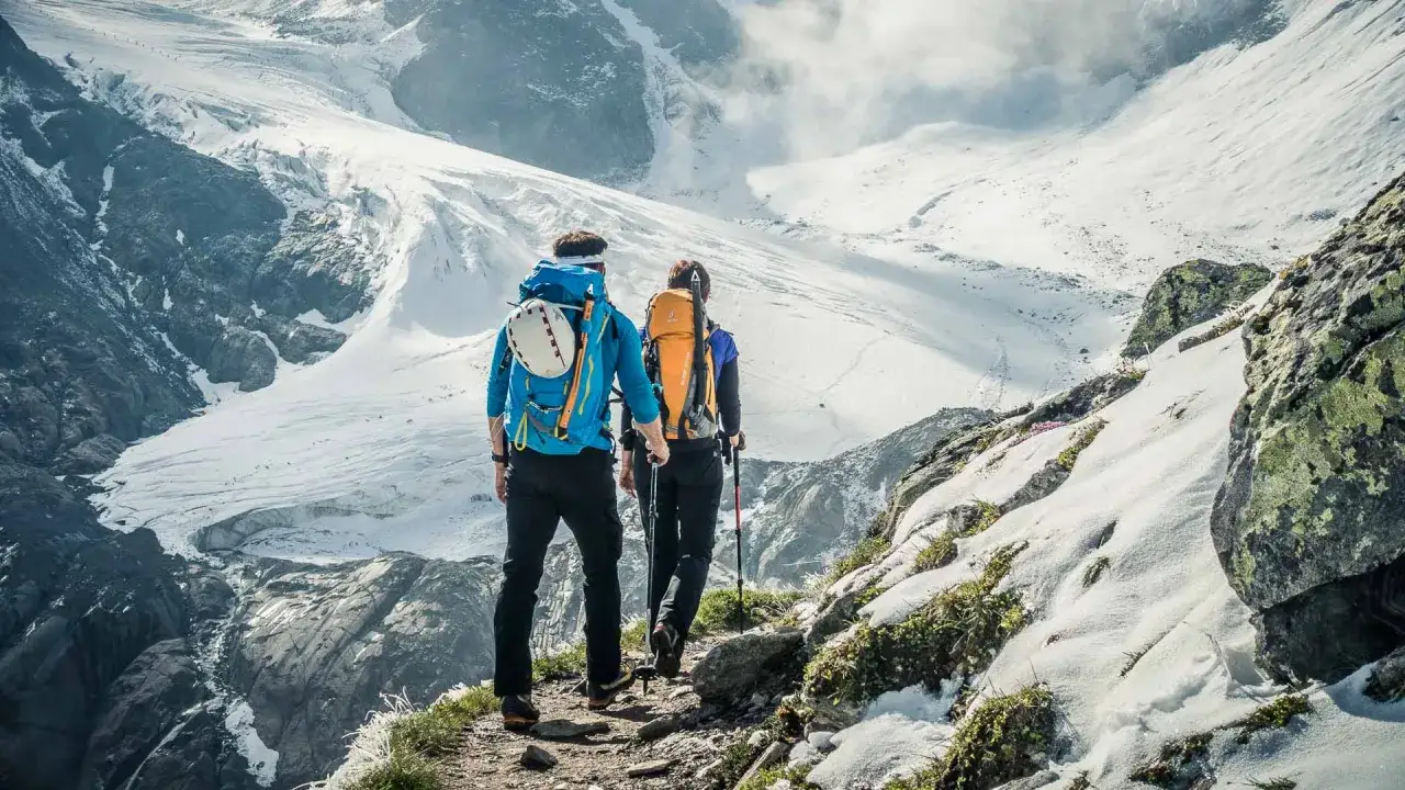 Zwei Bergsteiger auf dem Wildspitze Normalweg, mit Rucks&auml;cken und St&ouml;cken, steigen einen steinigen Pfad hinauf. Im Hintergrund erstreckt sich eine beeindruckende Gletscherlandschaft.