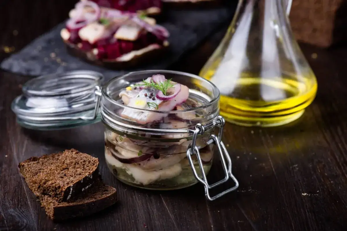Inlagd sill i glasburk med r&ouml;dl&ouml;k och dill. M&ouml;rkt br&ouml;d och en karaff med olja i bakgrunden. En klassisk svensk sillr&auml;tt.