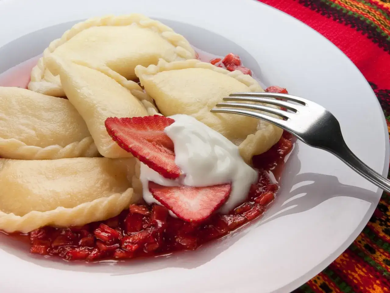 Pyszne pierogi z serem, polane sosem truskawkowym i udekorowane bitą śmietaną. Widoczny widelec zaprasza do spróbowania.