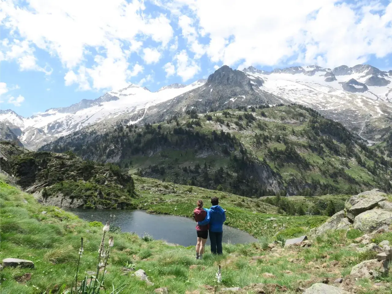 Pareja admira los ibones de Villamuerta, un lago de montaña rodeado de prados verdes y picos nevados.