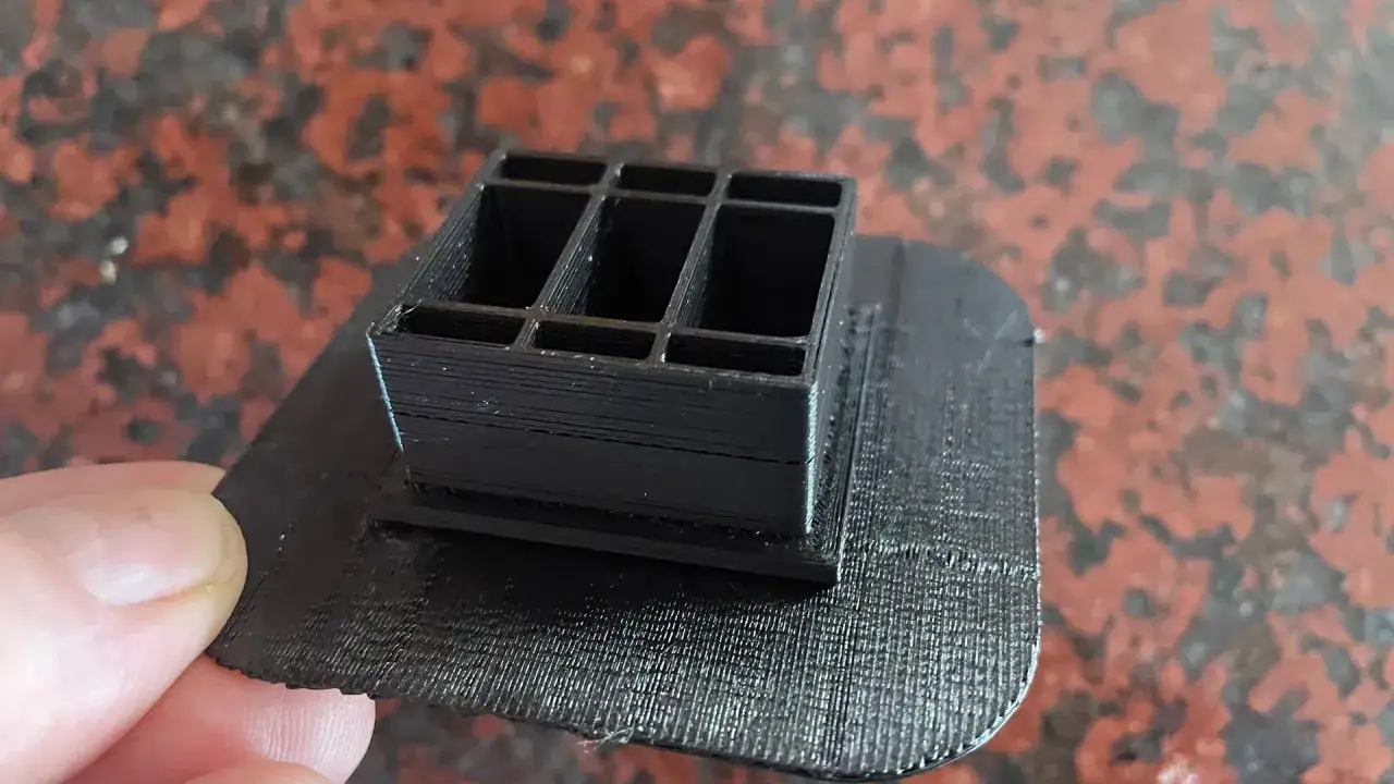 Czarny, wielokomorowy element wydrukowany w technologii 3D, osadzony na płaskiej podstawie. Widoczny raft druk 3d.