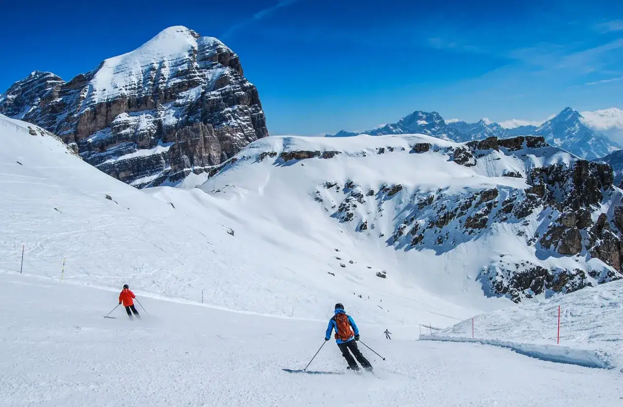 Narty w marcu na ośnieżonych stokach Dolomitów. Dwóch narciarzy zjeżdża po przygotowanej trasie, podziwiając majestatyczne, skaliste szczyty.