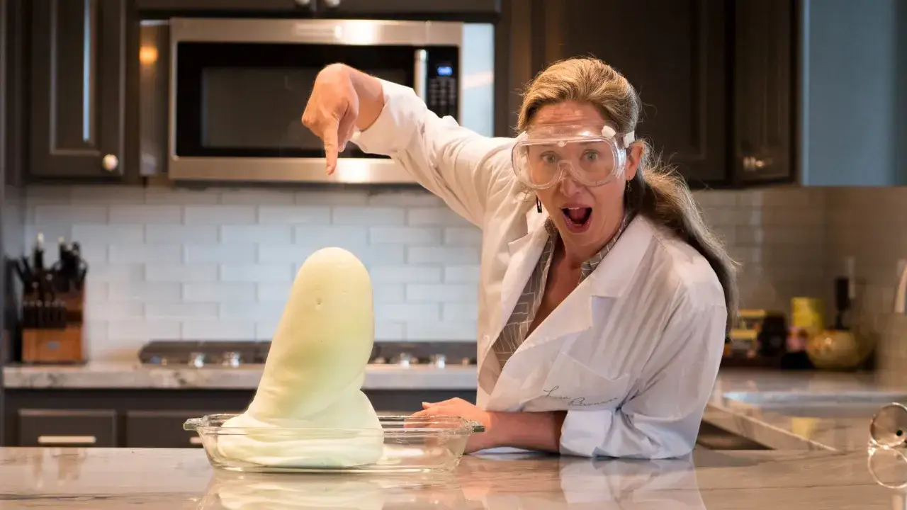 Frau in Laborkittel und Schutzbrille zeigt auf schäumende Masse. Ein spannendes Experiment zur Chemie im Alltag.