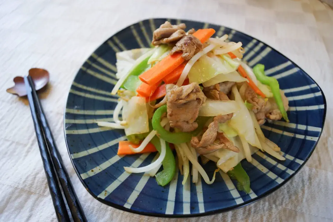 A delicious Japanese stir fry with tender pork, crisp cabbage, carrots, bean sprouts, and green peppers, served on a blue and white striped plate with chopsticks.