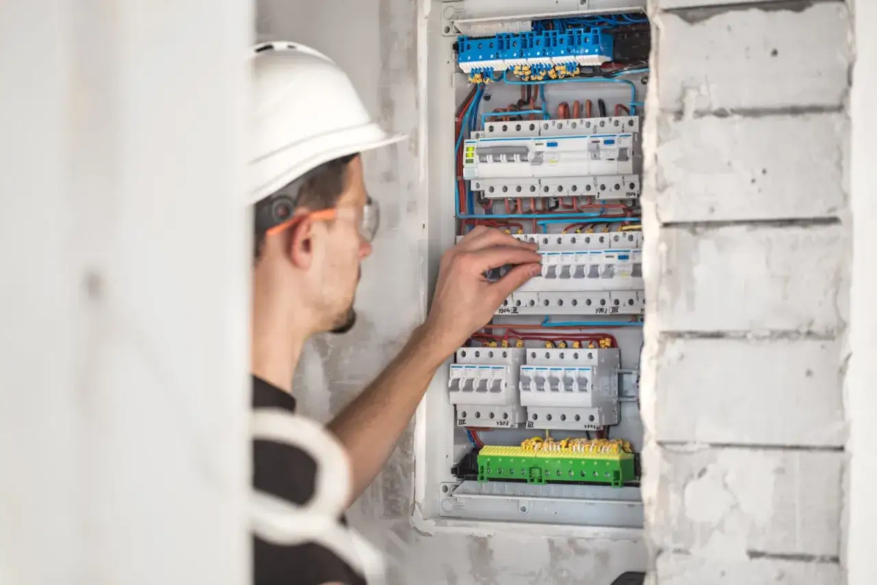 Elektryk w kasku i okularach ochronnych reguluje bezpieczniki w skrzynce. To ważny krok, by złożyć wniosek o obniżenie napięcia w sieci.