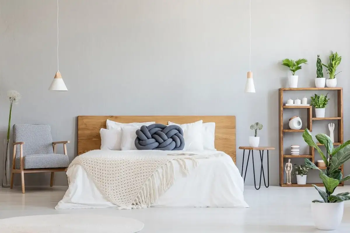 Camera da letto in stile nordico con letto bianco, coperta beige, cuscino grigio a nodo, poltrona a scacchi e scaffale in legno con piante.