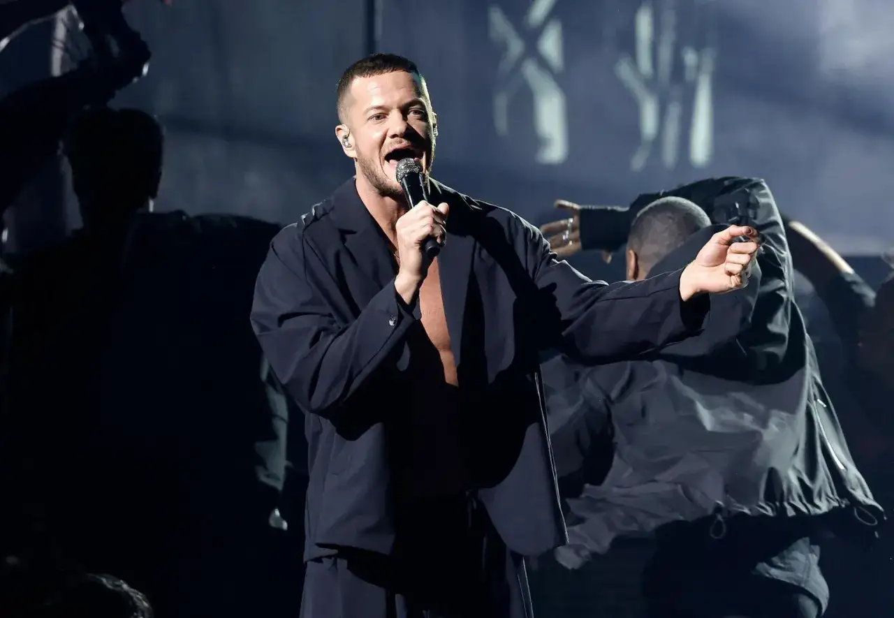 Imagine Dragons koncert Polska. Wokalista Dan Reynolds śpiewa do mikrofonu na scenie, otoczony tancerzami.