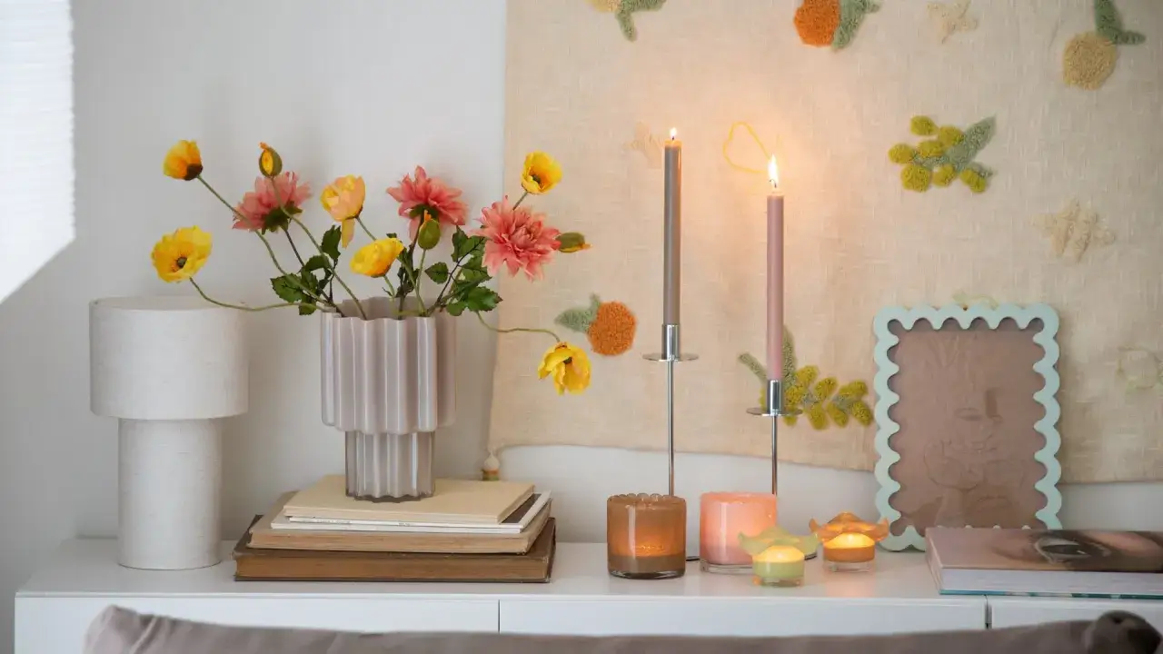 En mysig h&ouml;rna med blommor i en vas, ljus och en v&auml;ggbonad som bidrar till v&aring;r inredning.