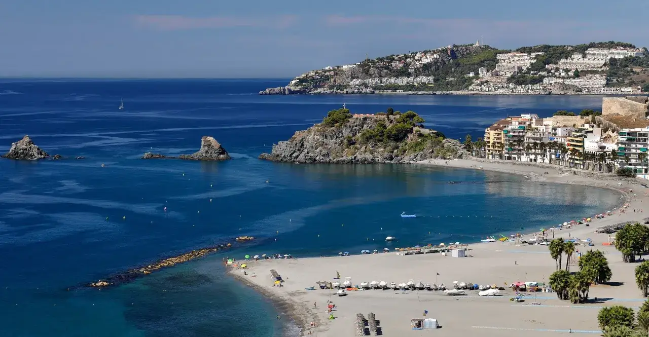 Bahía con pueblos de Granada con playa, rocas y barcos. Casas blancas se asoman a la costa.