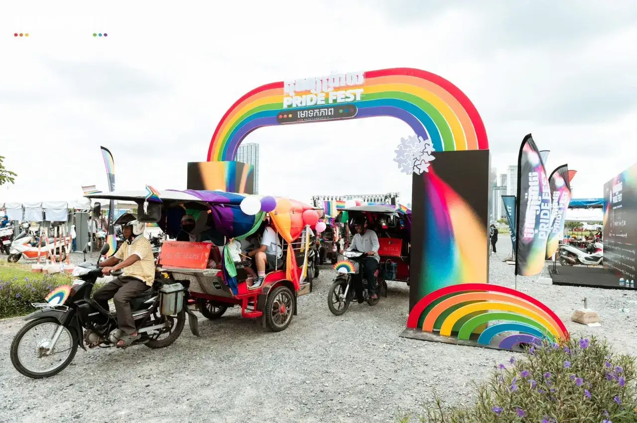 Tuk-tuks decorated with rainbow colors drive through a Pride Fest archway in Cambodia, celebrating gay pride.