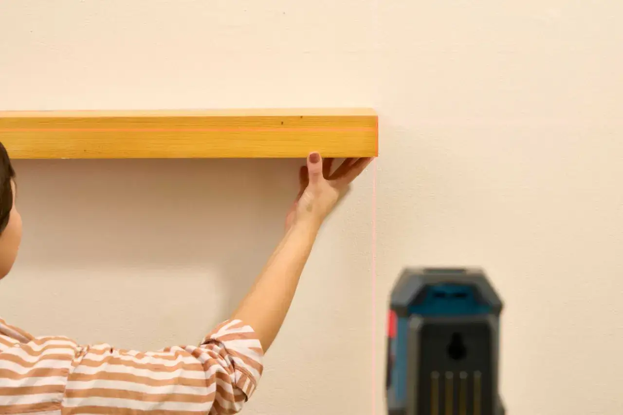 Une main ajuste une étagère en bois, guidée par les lignes roses d'un niveau laser pour une pose parfaite.