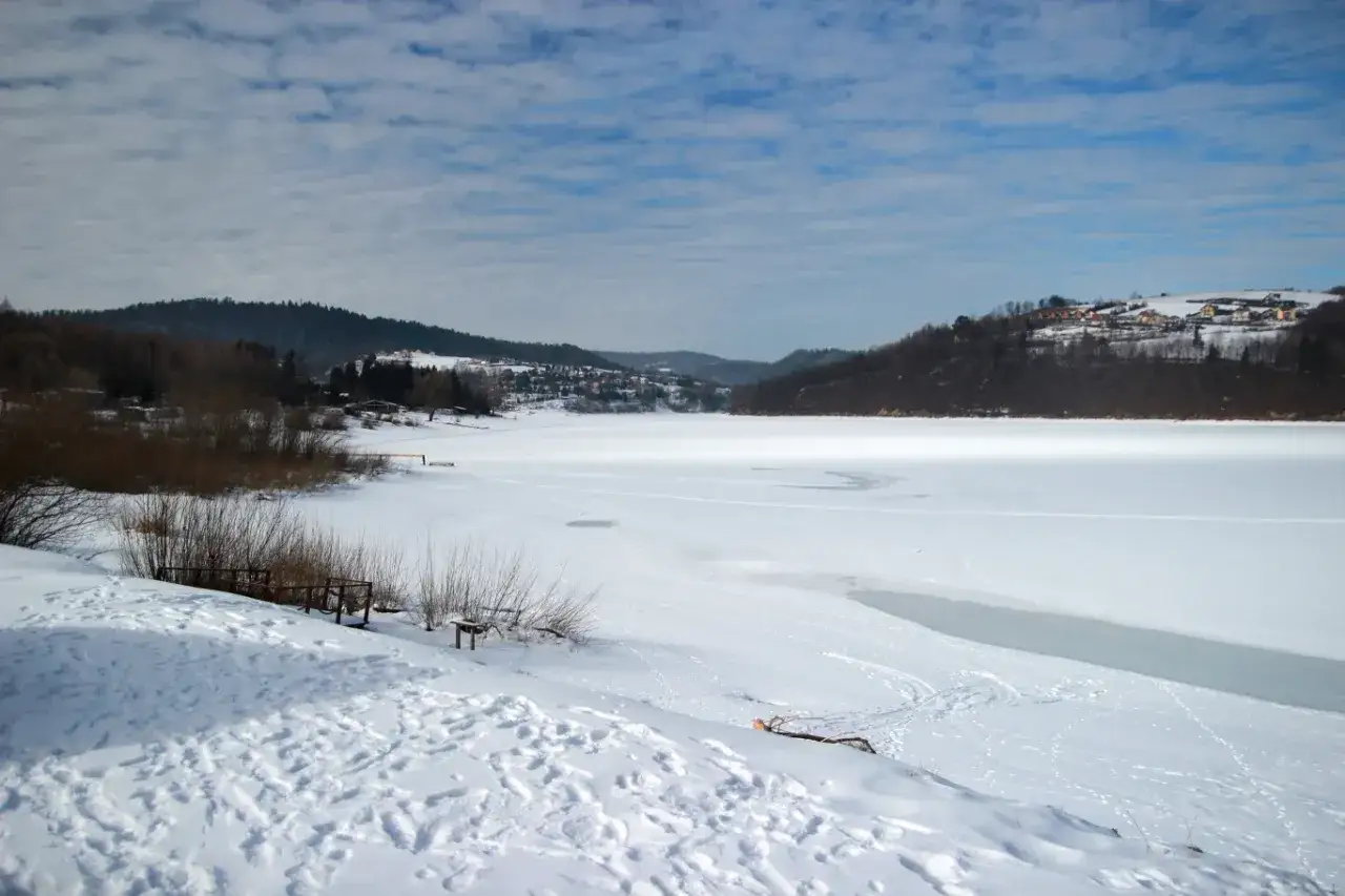 Zamarznięte jezioro Solina zimą. Ślady na śniegu i lodzie, drzewa, wzgórza z domkami w tle. Atrakcje zimowe w Bieszczadach.
