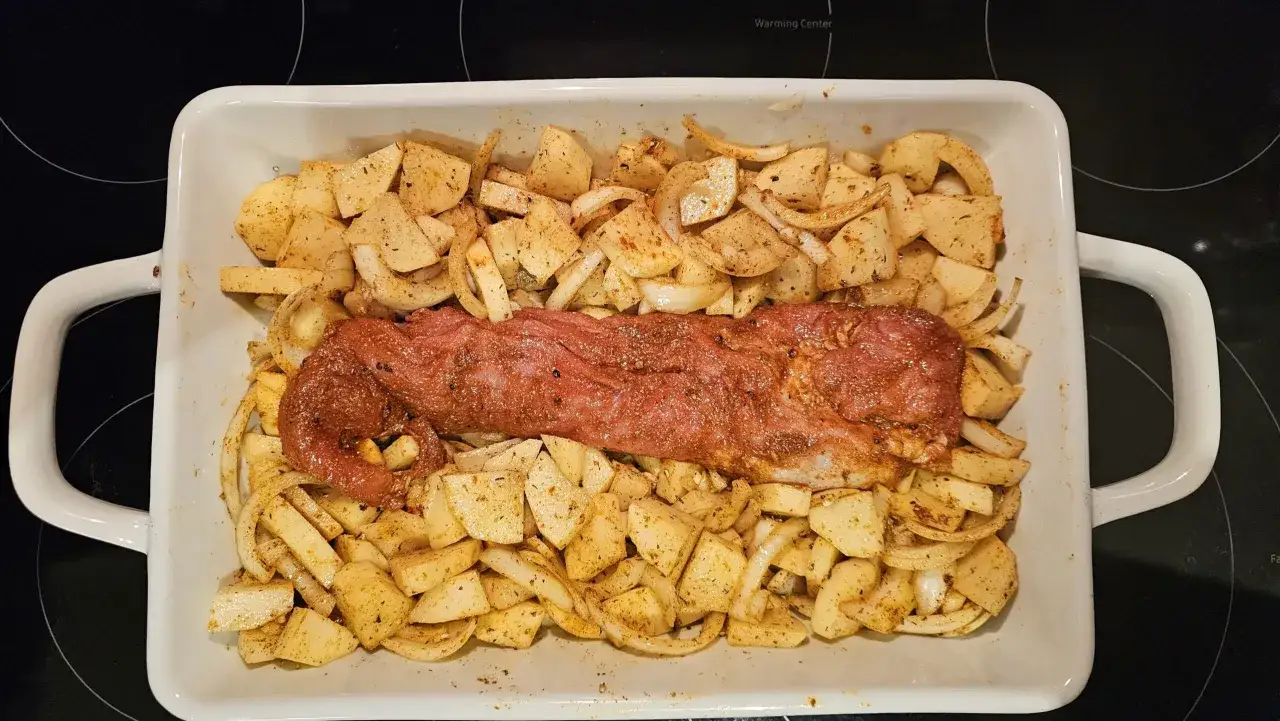 Filet mignon assaisonné, prêt à être rôti avec des pommes de terre et des oignons coupés en dés dans un plat blanc.