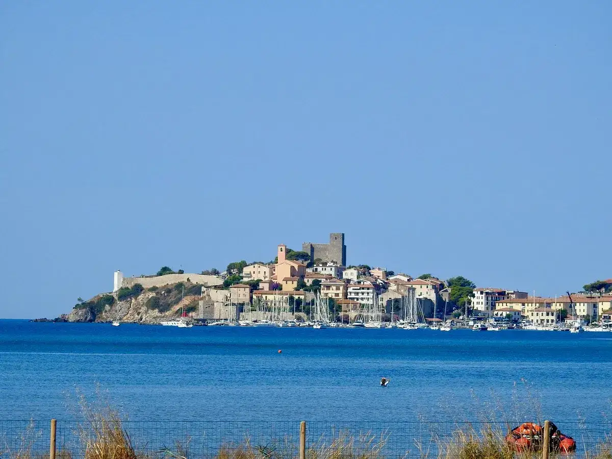 Il **Talamone centro storico** si affaccia sul mare blu, con un castello e case arroccate sulla collina. Barche a vela nel porto.