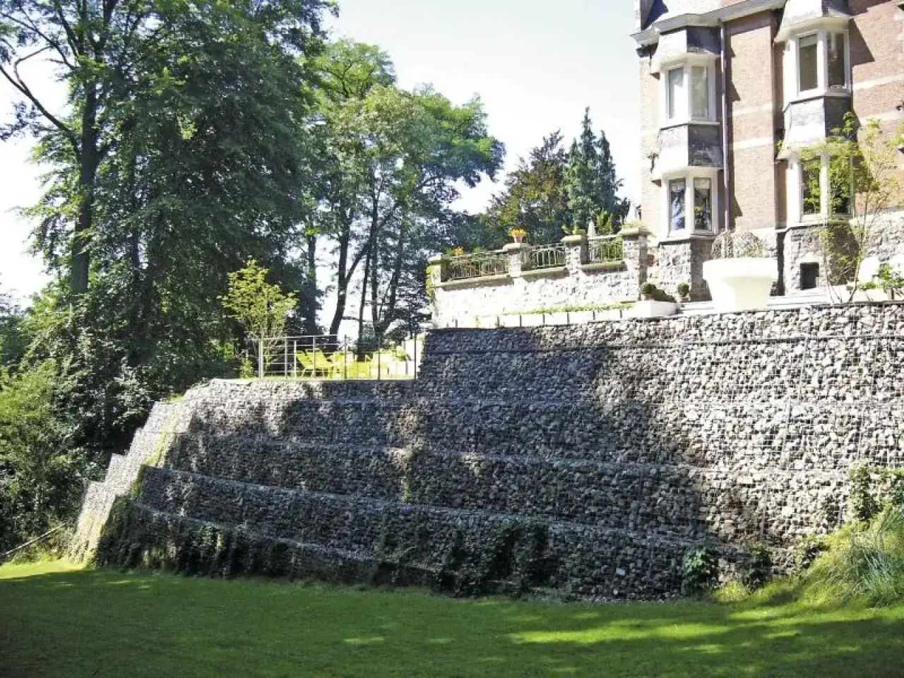 Imponująca skarpa na granicy działki, zbudowana z kamieni w gabionach, tworzy tarasowy krajobraz. Na g&oacute;rze widać fragment rezydencji.