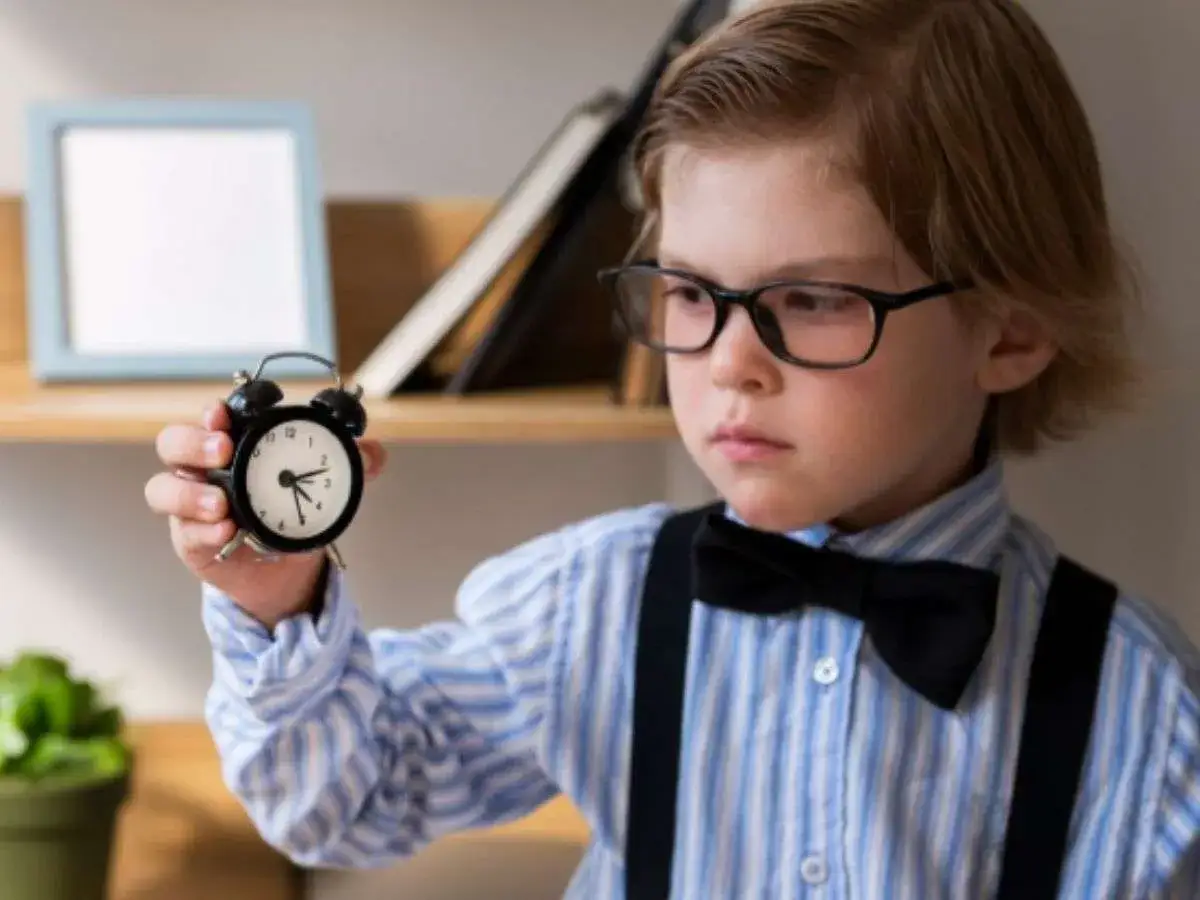 Niño con gafas y pajarita sostiene un despertador. Son las horas en punto y media, ¡hora de aprender!