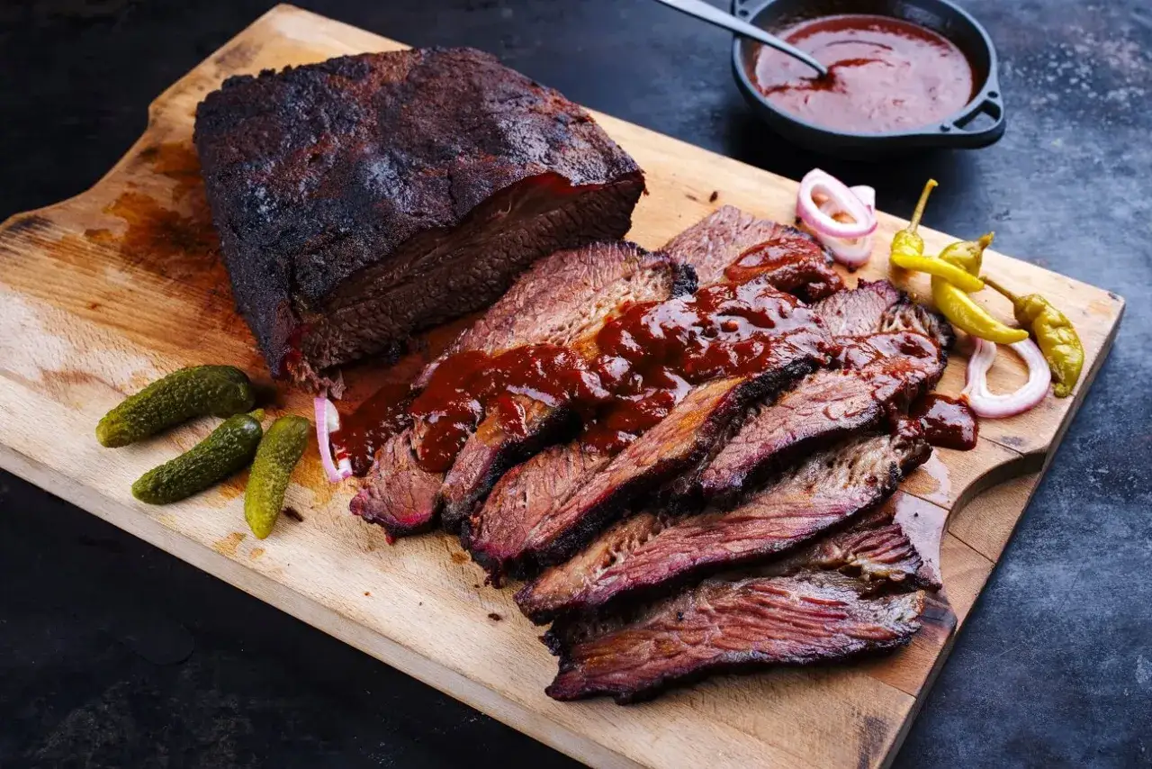Saftiges, aufgeschnittenes pulled pork mit BBQ-Sauce, Cornichons und eingelegten Zwiebeln auf einem Holzbrett.