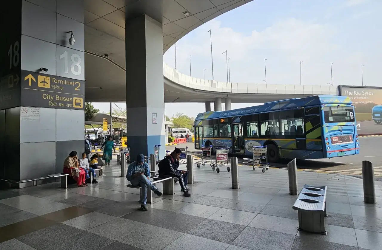 Oczekujący na autobusy na lotnisku Delhi. W tle niebieski autobus elektryczny.