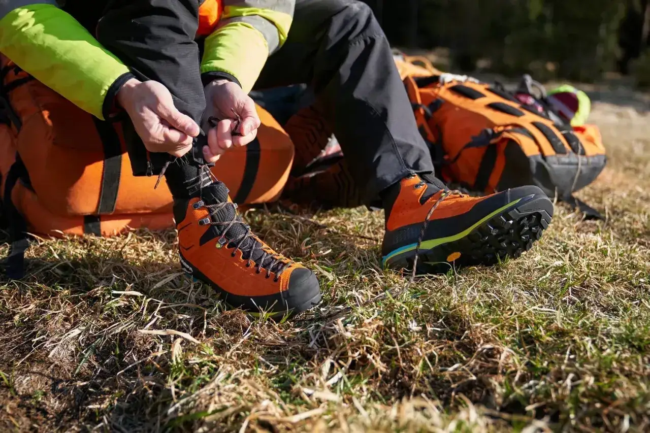 Osoba zawiązuje sznur&oacute;wki pomarańczowych but&oacute;w trekkingowych, kt&oacute;re obcierają pięty. Obok leży plecak.