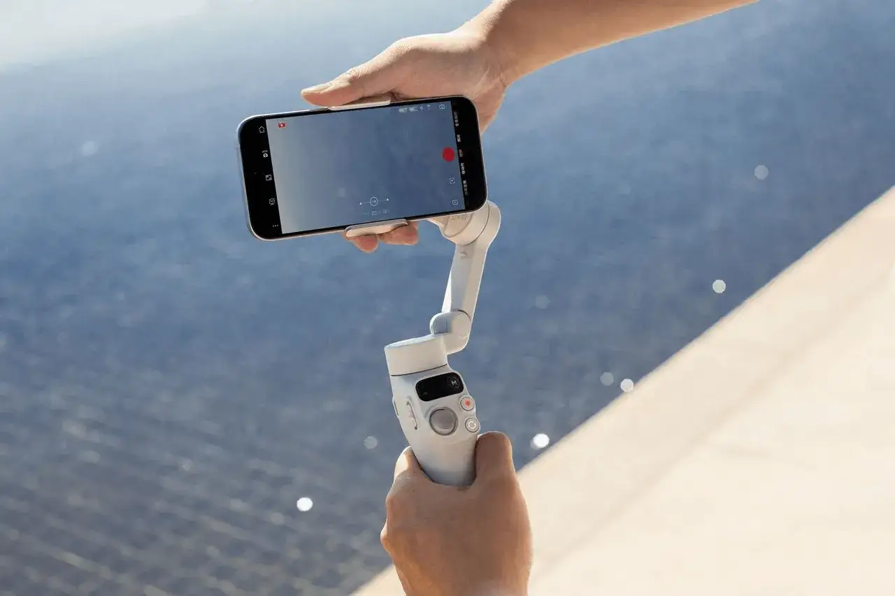 Ręce trzymają smartfon zamontowany na białym gimbala, gotowe do nagrywania płynnych ujęć.