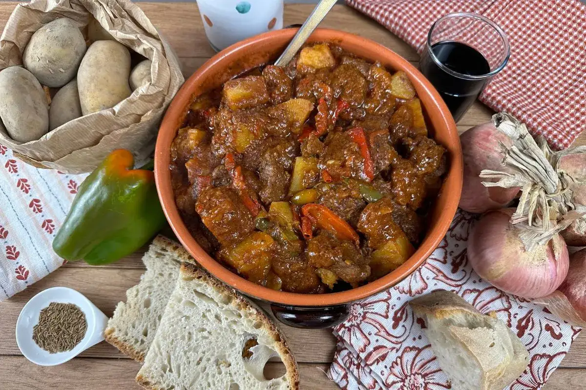 Un saporito gulash di manzo fumante in una ciotola di terracotta, accompagnato da patate, peperoni, cipolle, pane e spezie.