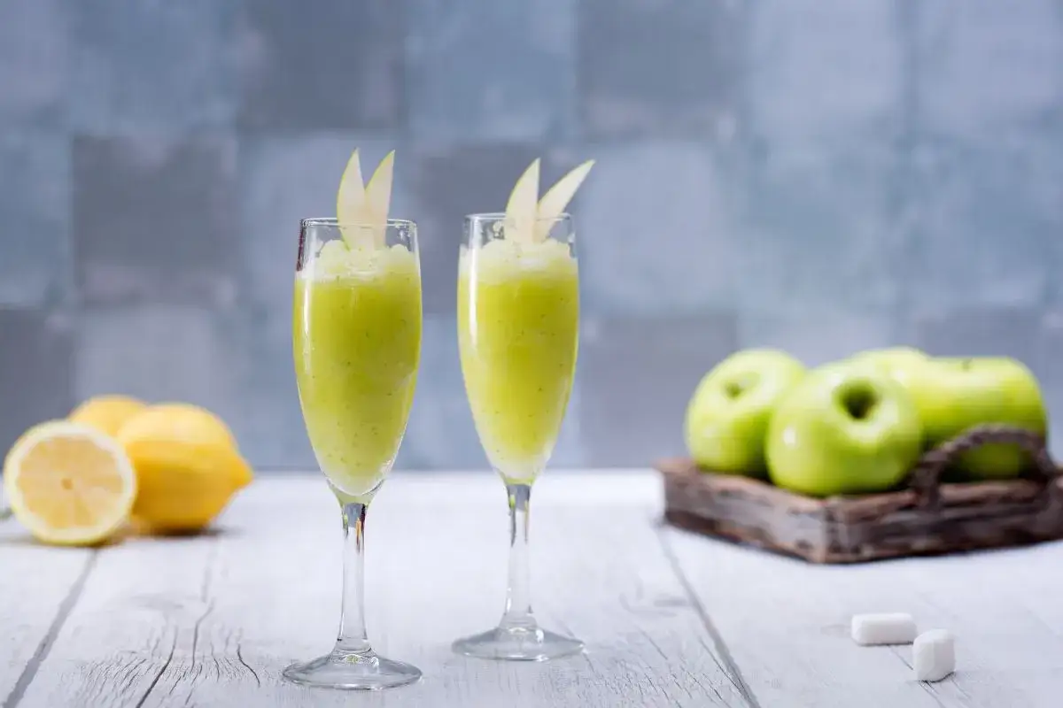 Zwei Gläser mit erfrischendem Apfel Sorbet, garniert mit Apfelscheiben. Im Hintergrund grüne Äpfel und Zitronen.