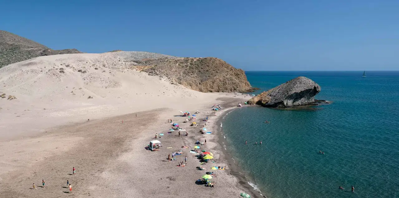 Sonnige Playa de M&oacute;nsul mit Sandd&uuml;nen, Felsen und badenden Menschen im t&uuml;rkisfarbenen Meer.