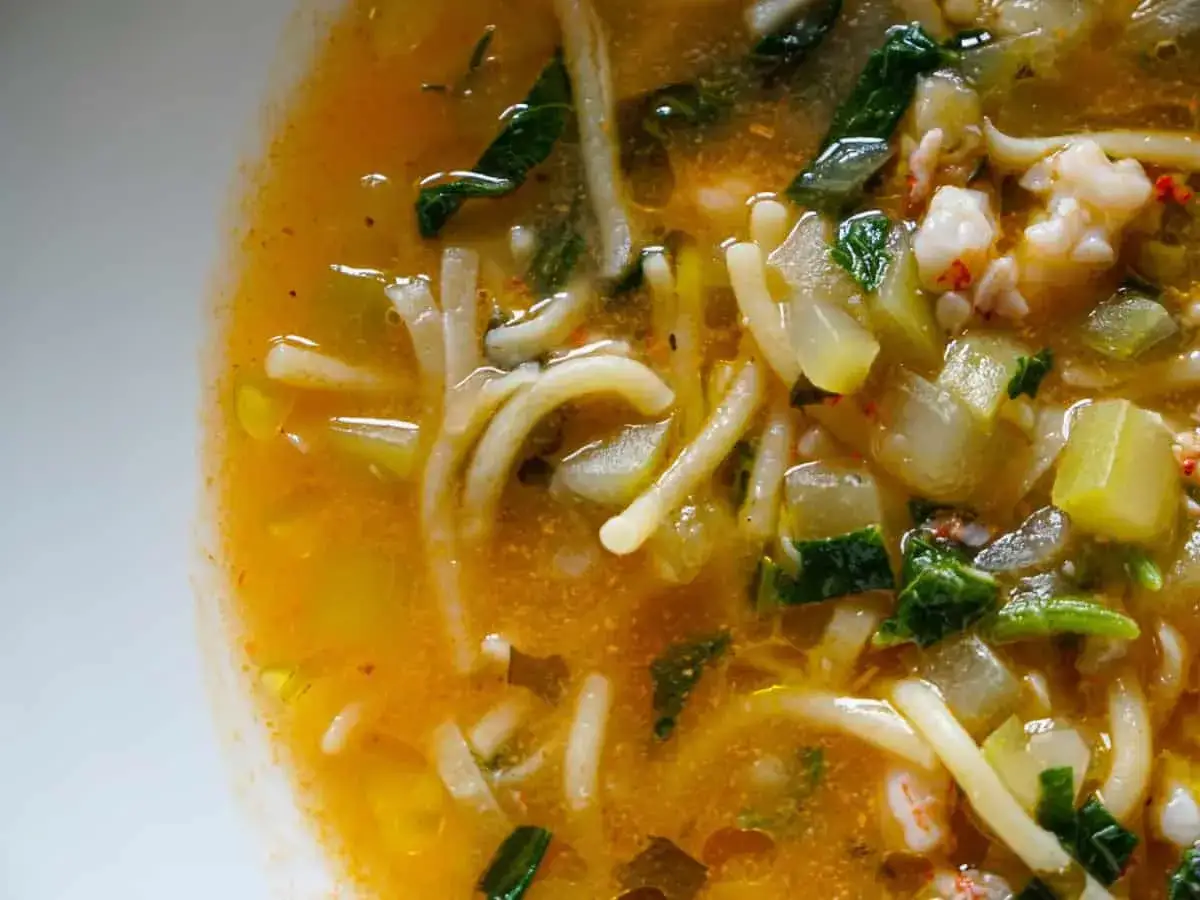 Primo piatto fumante di pasta con i tenerumi, verdure e piccoli gamberetti in un brodo dorato.