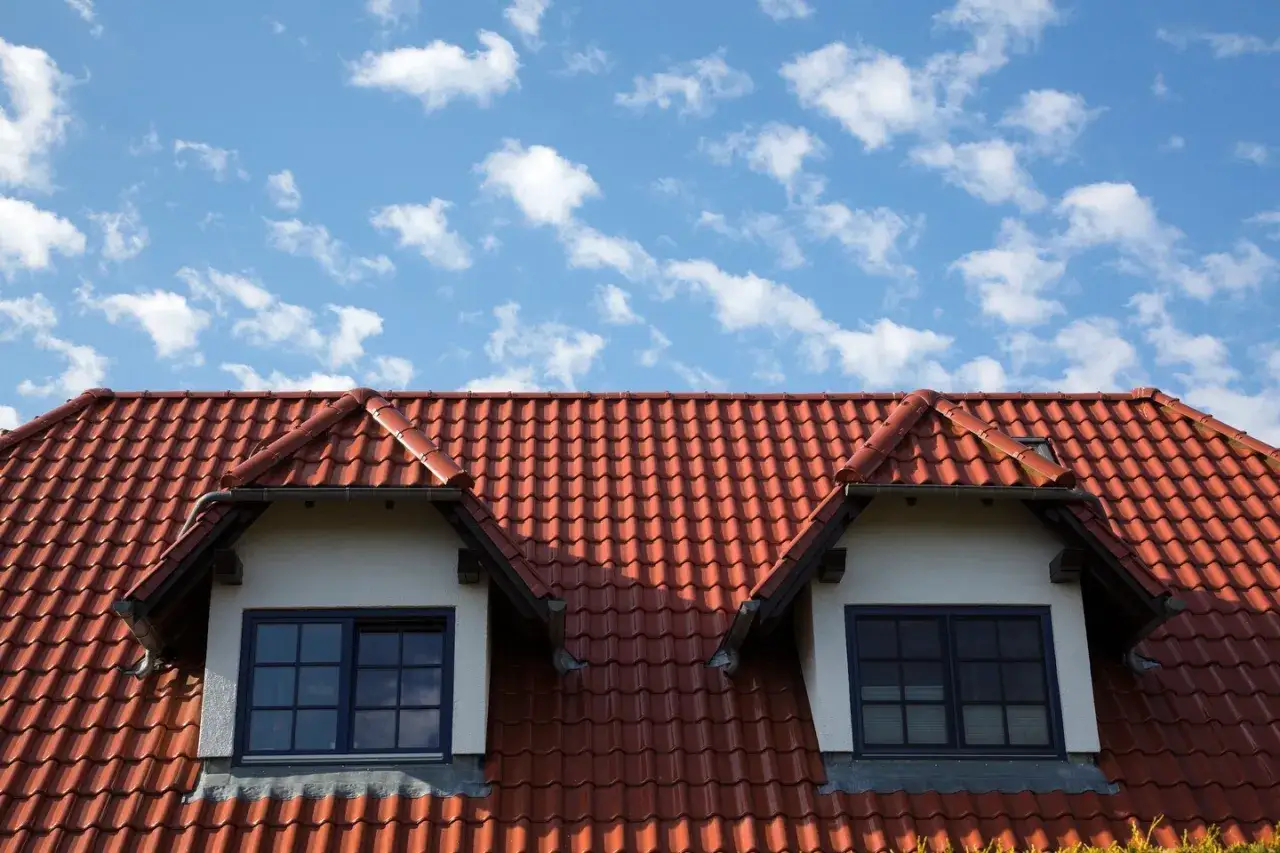 Dachówka ceramiczna w kolorze ceglastym pokrywa dach domu z dwoma lukarnami. Różne rodzaje dachówek tworzą falisty wzór.