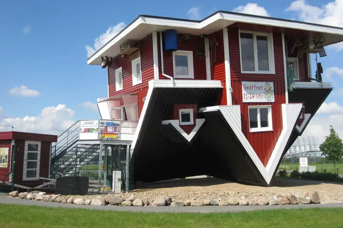Ein rotes **Haus auf dem Kopf** steht auf einem Kiesbett. Die Fenster und das Dach sind nach unten gerichtet, als ob es umgedreht wäre.