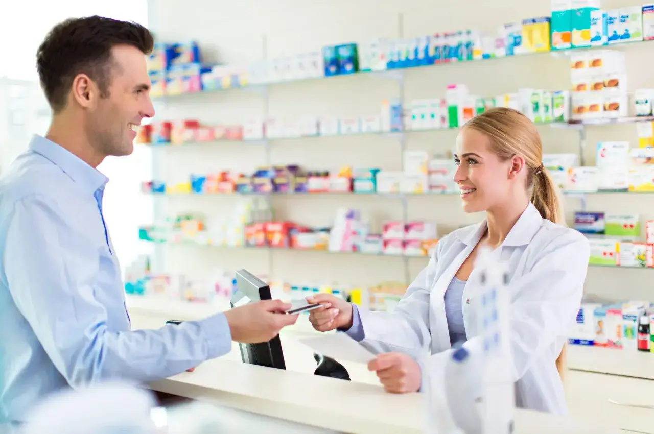 Uśmiechnięty klient płaci kartą, a technik farmacji obsługuje go przy ladzie aptecznej.