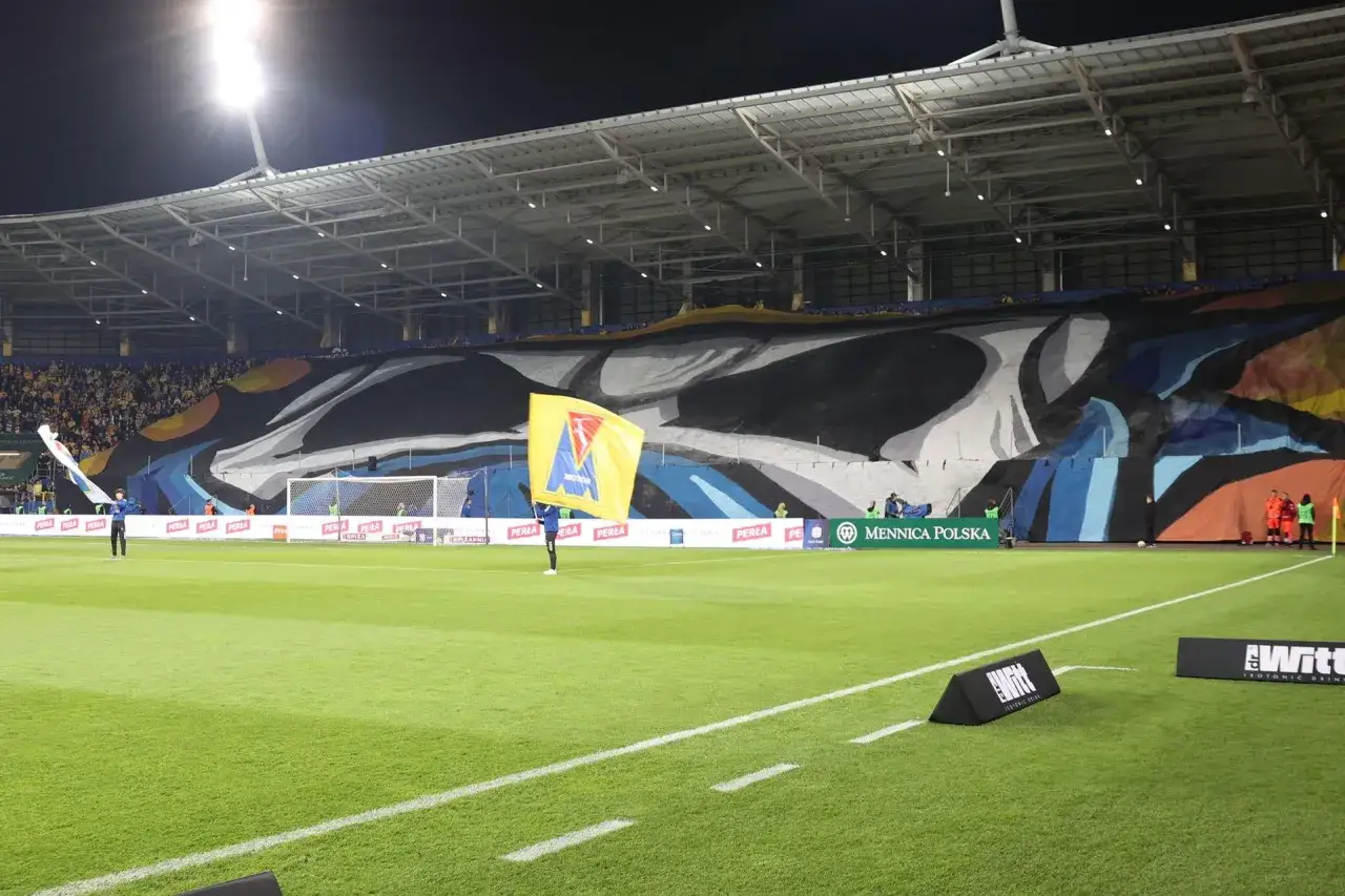 Wielka oprawa kibiców lubelskiego klubu piłkarskiego Motor Lublin na stadionie. Żółta flaga z herbem klubu powiewa na tle murawy i trybun.