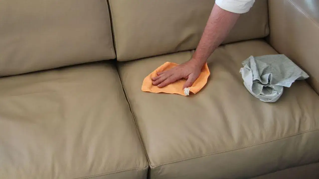 Mano che pulisce un divano in pelle beige con un panno arancione. Impara come togliere lo sporco dalla pelle per mantenerla come nuova.