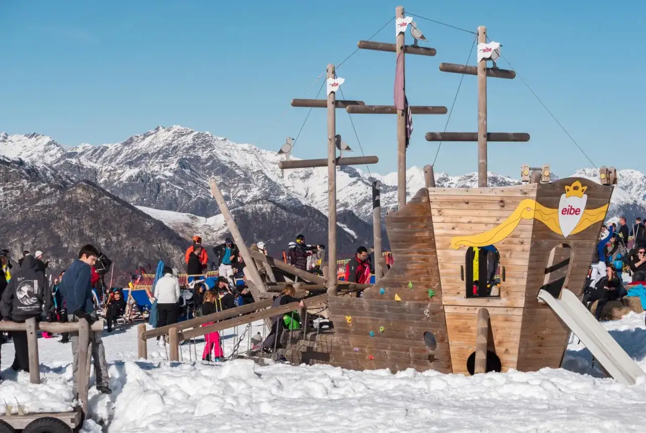 Un'area giochi a forma di nave pirata sulla neve, con montagne innevate sullo sfondo, all'Alpe di Mera. Bambini e adulti si divertono.