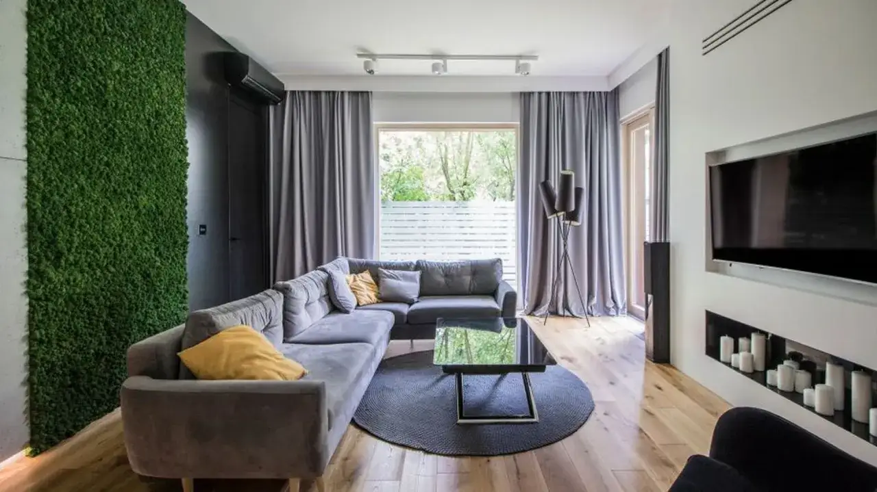 Sofa w kolorze popielatym, zielona ściana z mchu, czarny stolik i telewizor.