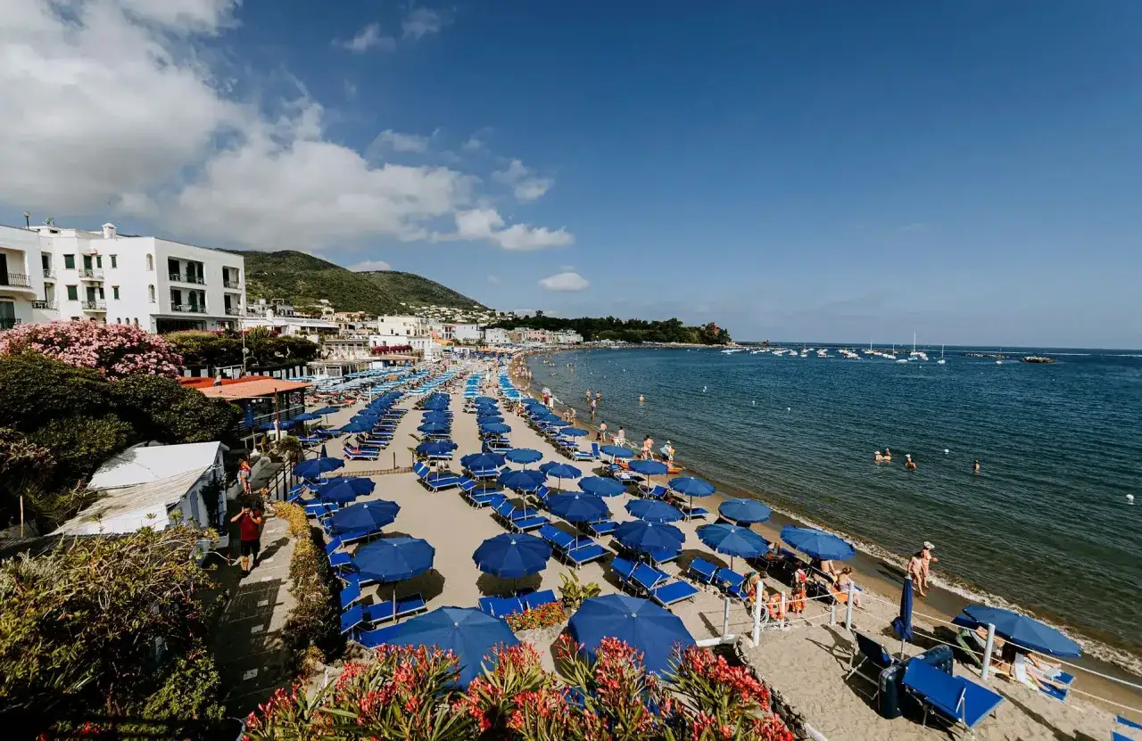 Spiaggia affollata con ombrelloni blu e lettini, Hotel Bristol Ischia sullo sfondo, mare azzurro e cielo limpido.