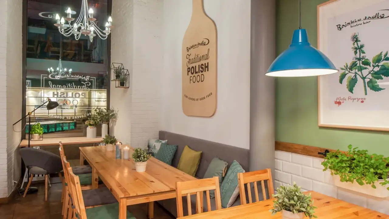 Przytulne wnętrze bistro i restauracji z drewnianymi stołami, wygodnymi kanapami i dekoracjami ściennymi promującymi tradycyjne polskie jedzenie.