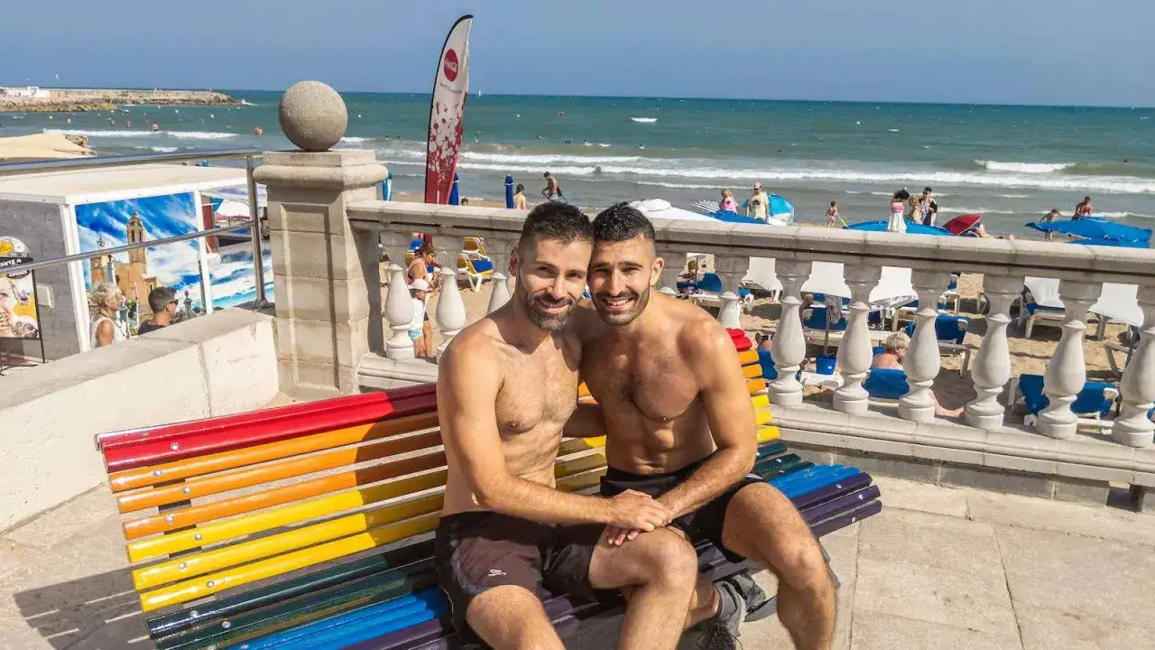 A happy couple enjoys a sunny day at the Sitges gay beach, sitting on a rainbow bench with the ocean in the background.