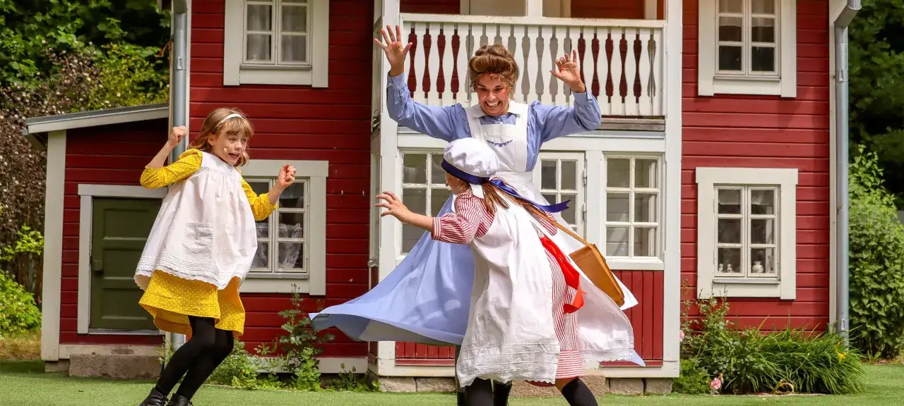 Barn leker vid Junibackens Madicken-hus. En vuxen i blå klänning och vit förkläde, två flickor i klänningar.