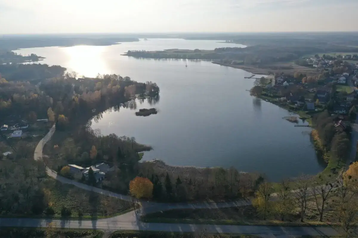 Jesioro Kałębie o poranku, otoczone jesiennymi drzewami i domami. Słońce odbija się od spokojnej tafli wody.