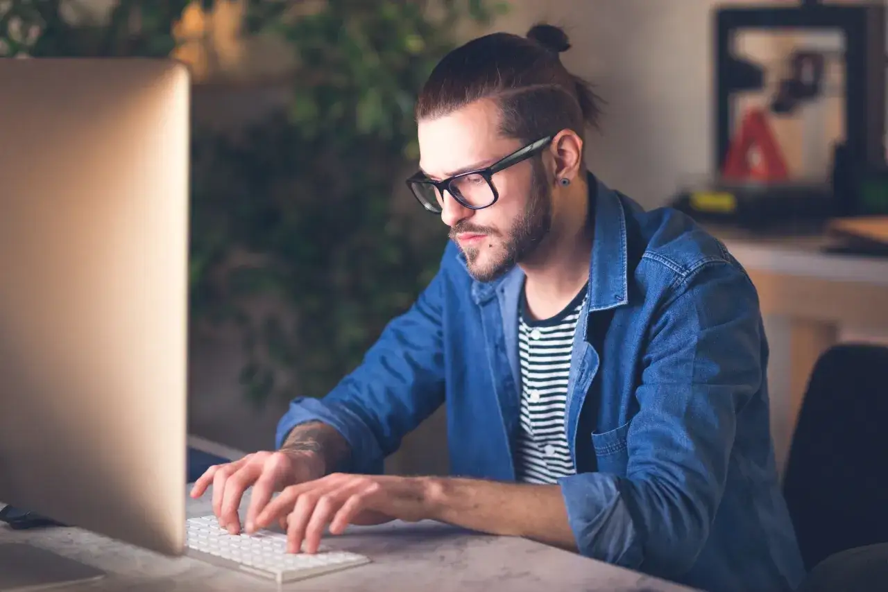 Giovane uomo con occhiali e coda di cavallo, concentrato sul suo lavoro da giornalista al computer.