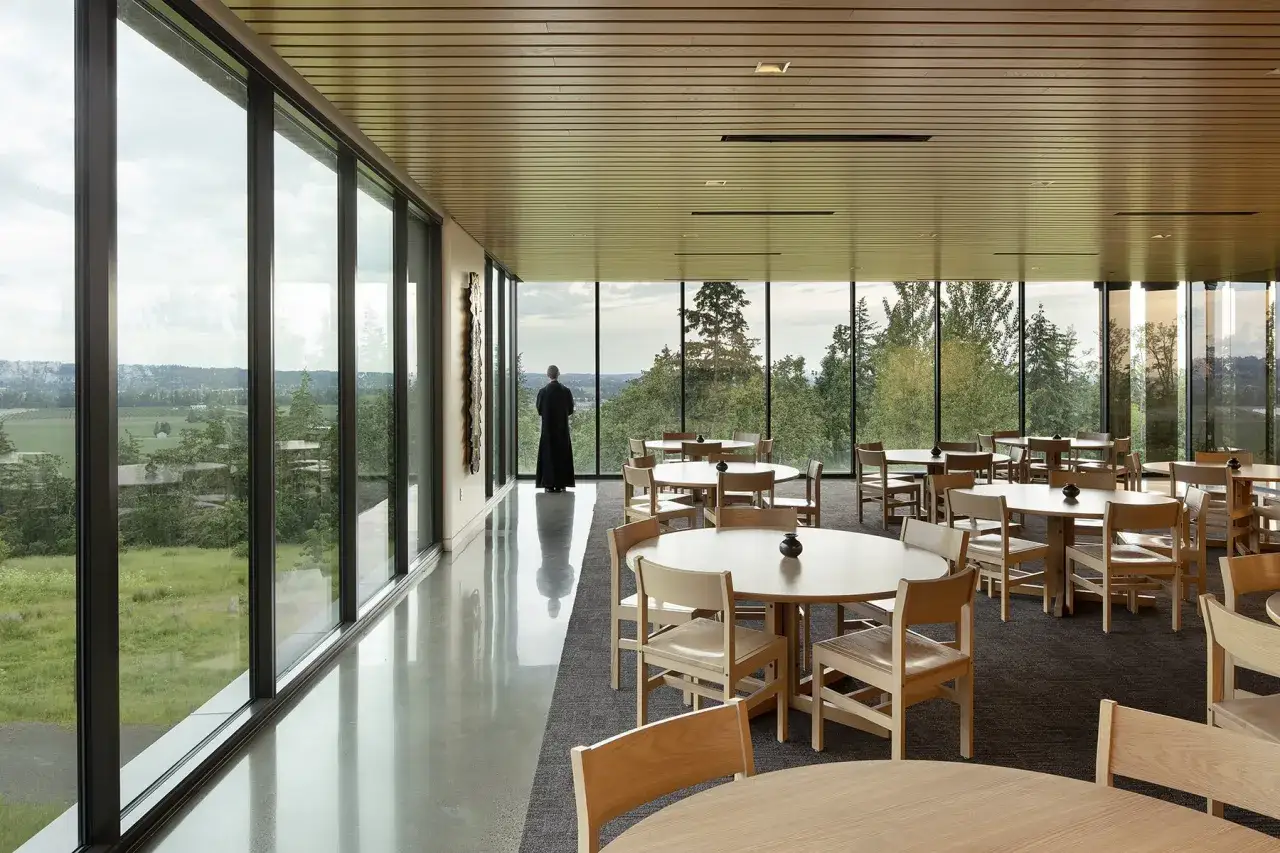 A monk gazes out at the rolling hills from the abbey guest house, a serene dining room with wooden tables and chairs.