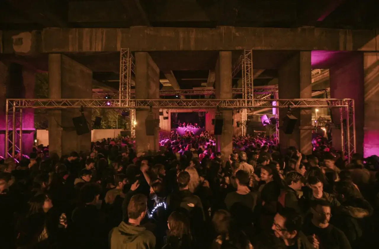 Foule dansante sous un pont, ambiance électrique pour le virage paris. Lumières roses et scène au loin.